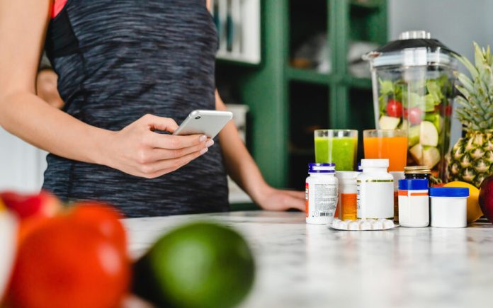 Young woman searching info about food supplements on her phone with fruits and additives on the table